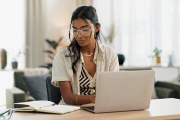 A woman in gold wire rimmed glasses sits at a laptop typing and preparing a resume using ChatGPT.